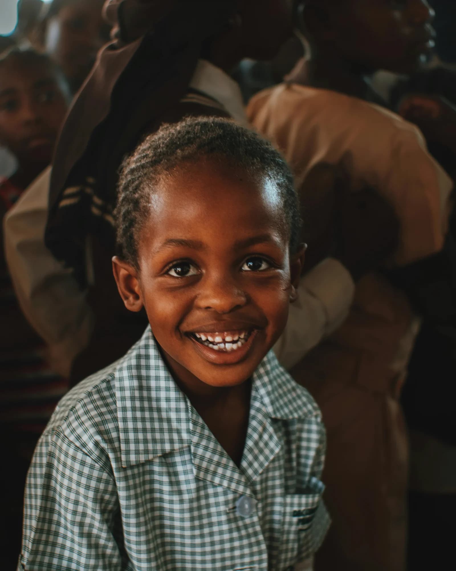 A smiling schoolchild in a checked uniform among a gathered crowd.